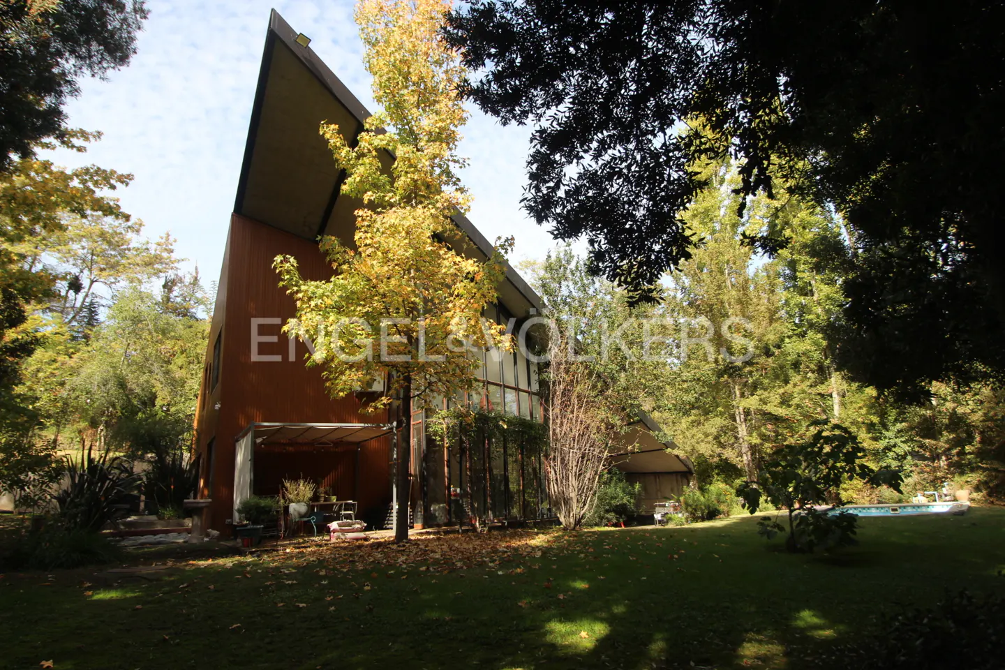 Modern brown house with a sharply angled roof, surrounded by trees and a green lawn. A pool is visible in the background.