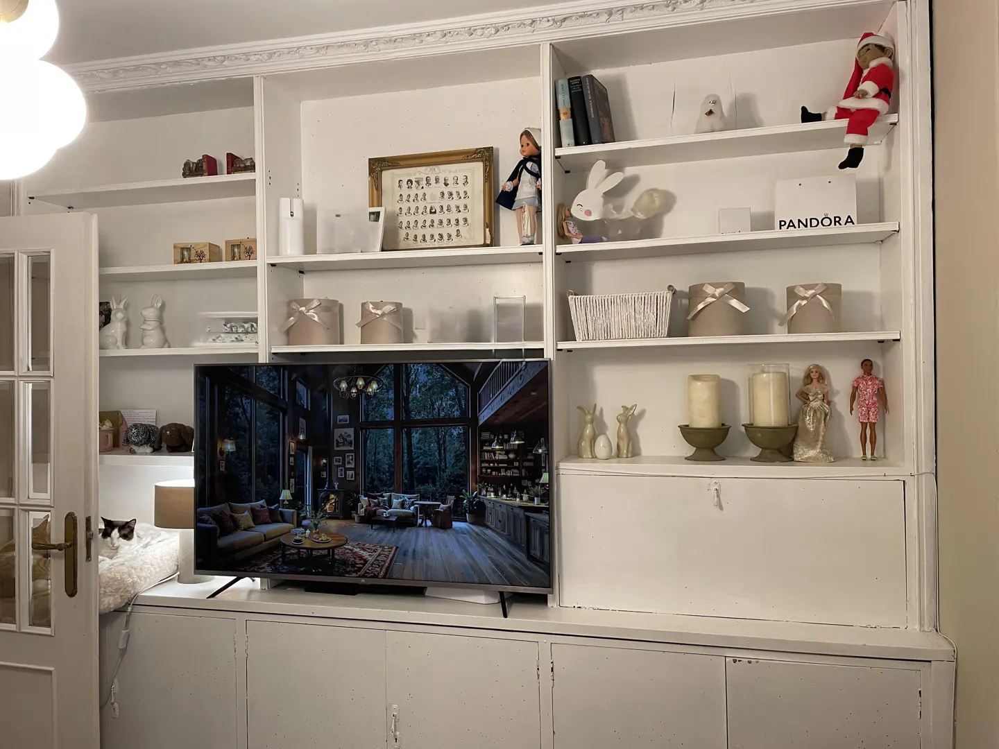White built-in shelving unit with decorative items and a TV displaying a living room scene. A cat rests in a bed to the left.
