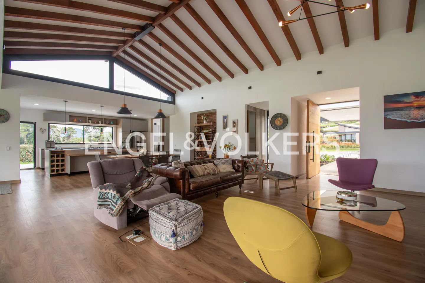 Open-concept living room with wood floors, exposed beams, and a mix of modern and rustic furniture. Large windows offer outdoor views.