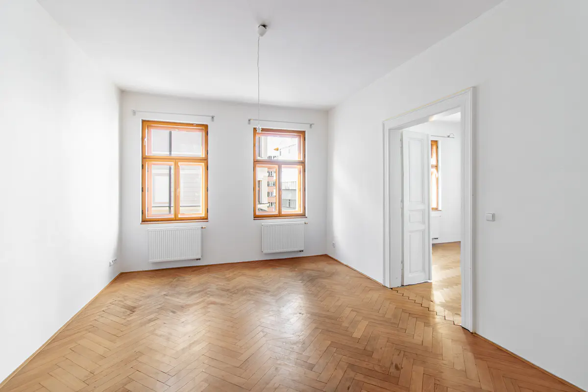 Bright, empty room with white walls, herringbone wood floor, two windows with wood frames, and a white doorway leading to another room.