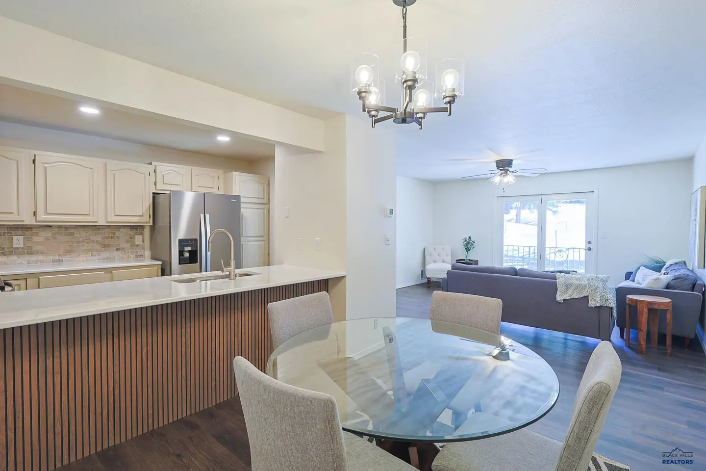 Open-concept kitchen and living room with a glass table, gray chairs, and a modern chandelier. Stainless steel appliances and dark wood floors.