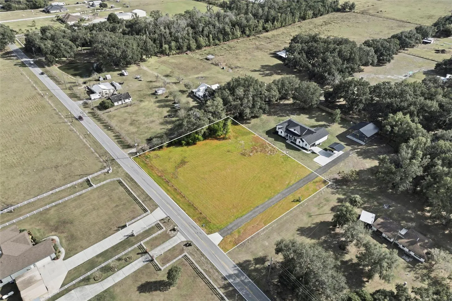 Aerial view of a vacant rectangular lot outlined in white, situated next to a road and a modern farmhouse in a rural area.