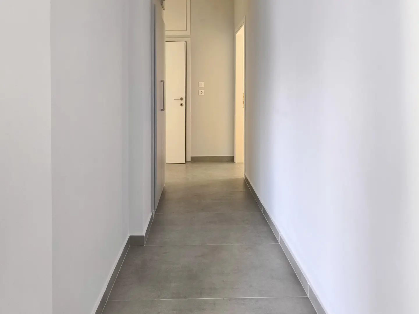A bright, minimalist hallway with white walls, grey tiled flooring, and closed white doors leading to other rooms.