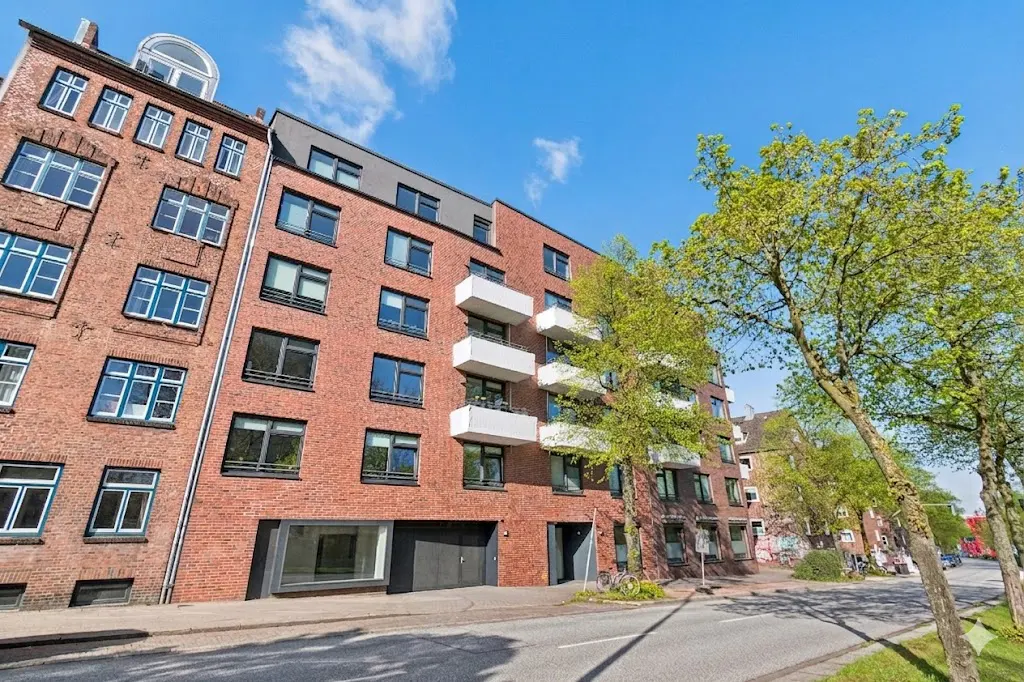 Exterior view of a red brick apartment building with white balconies and green trees under a blue sky.