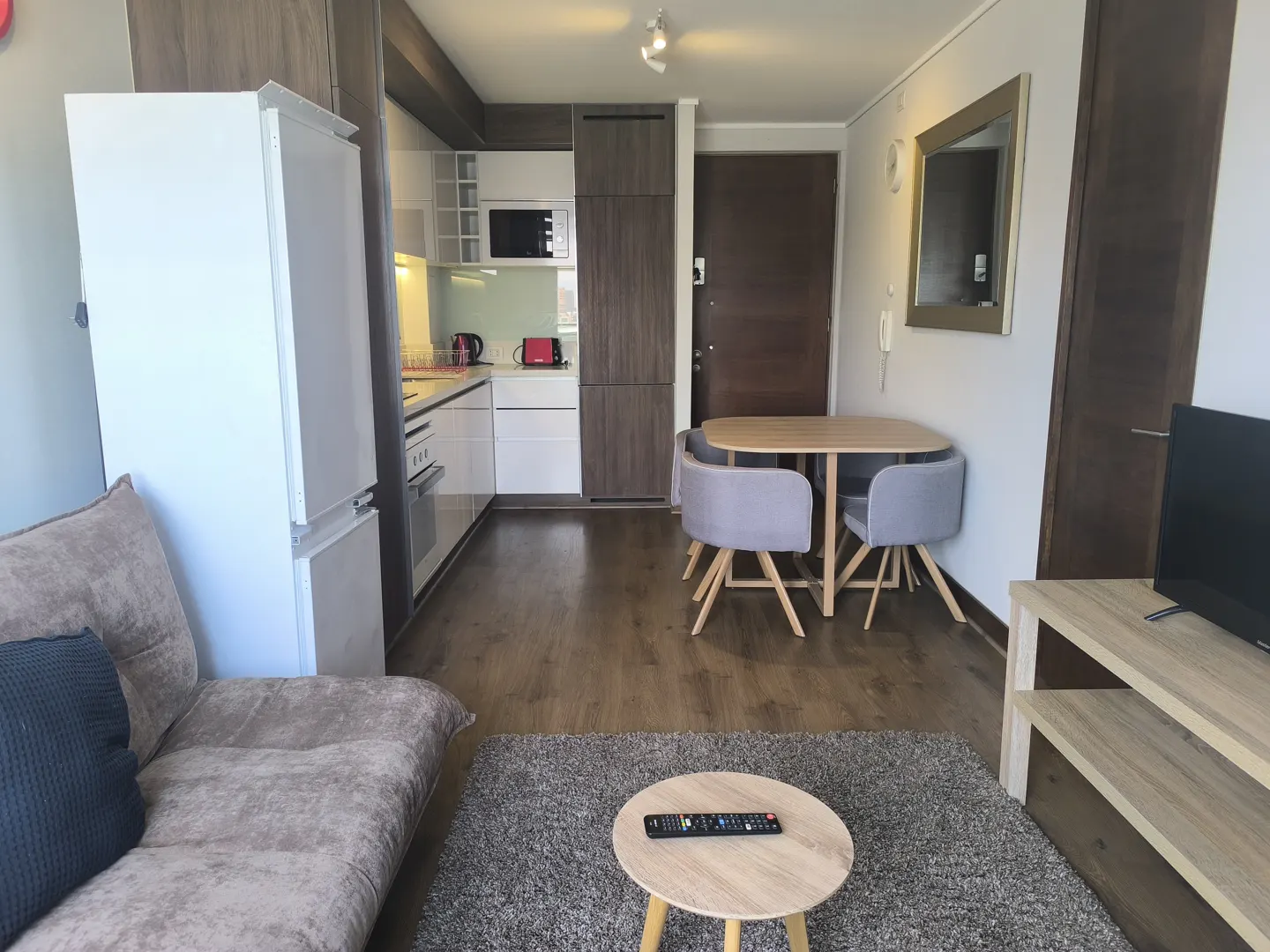 Interior view of a compact apartment with a kitchen, dining area, and living space. A sofa, table, and TV stand furnish the living area.