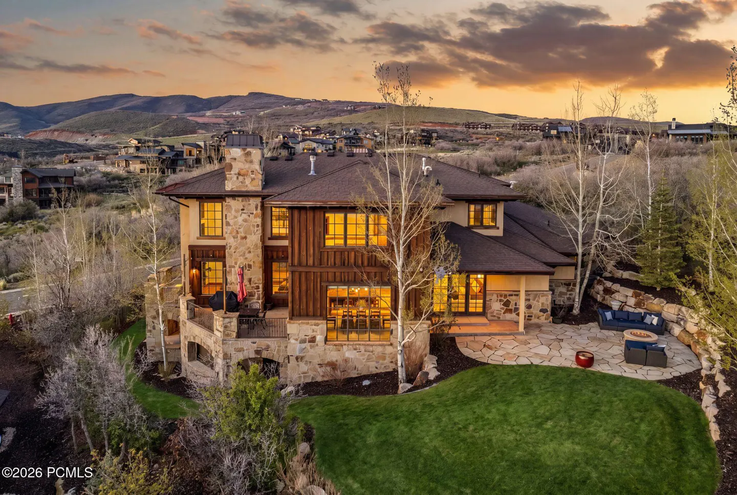 Exterior view of a large, multi-story home with stone and wood siding at sunset. Patio with outdoor seating and a green lawn.
