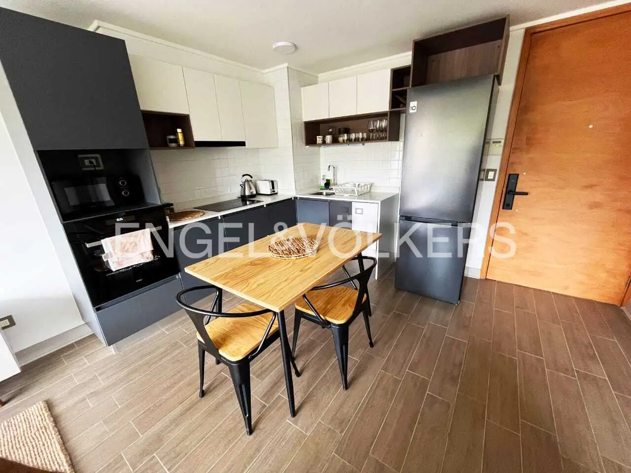 A modern kitchen with white cabinets, gray lower cabinets, and a wooden table with two black chairs. A stainless steel refrigerator stands near a wooden door.
