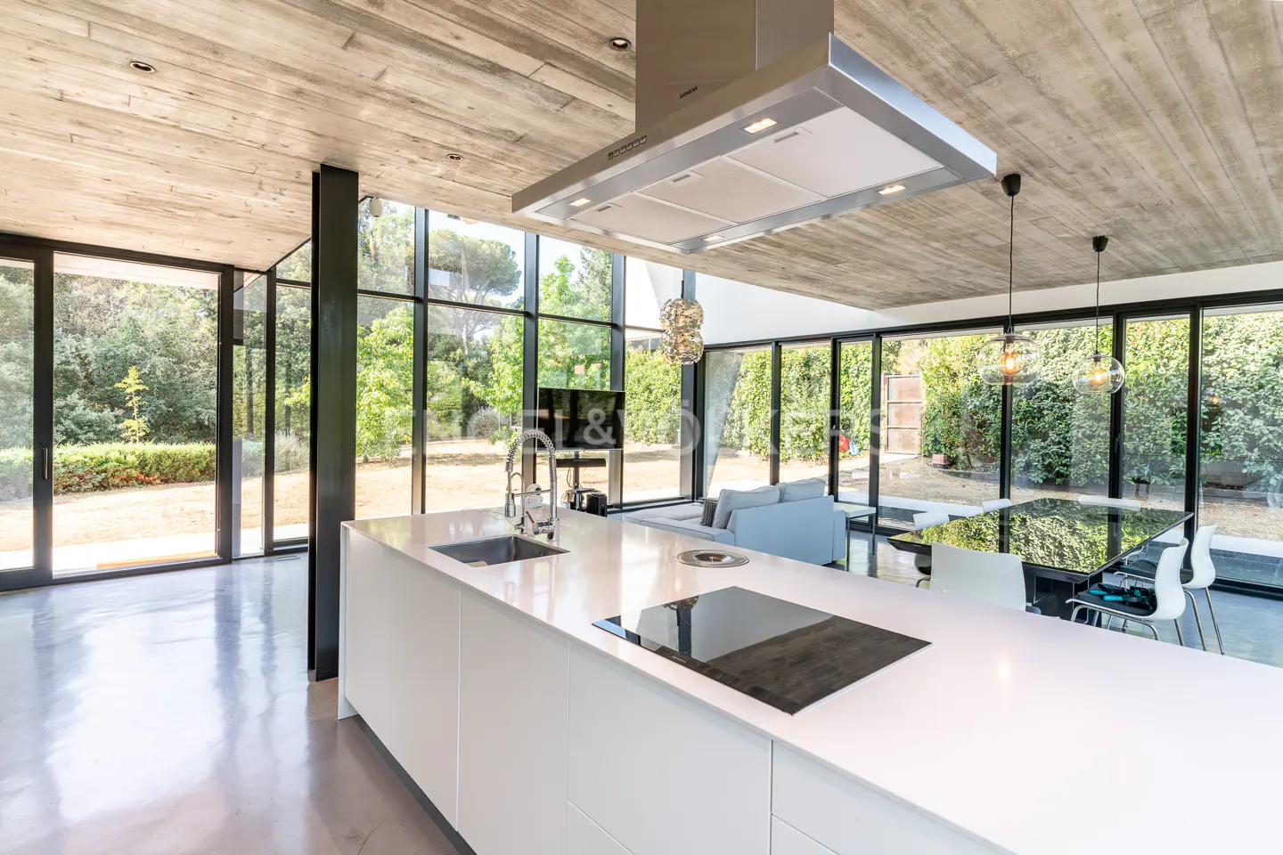 A modern kitchen with a white island, stainless steel range hood, and floor-to-ceiling windows overlooking a green yard.