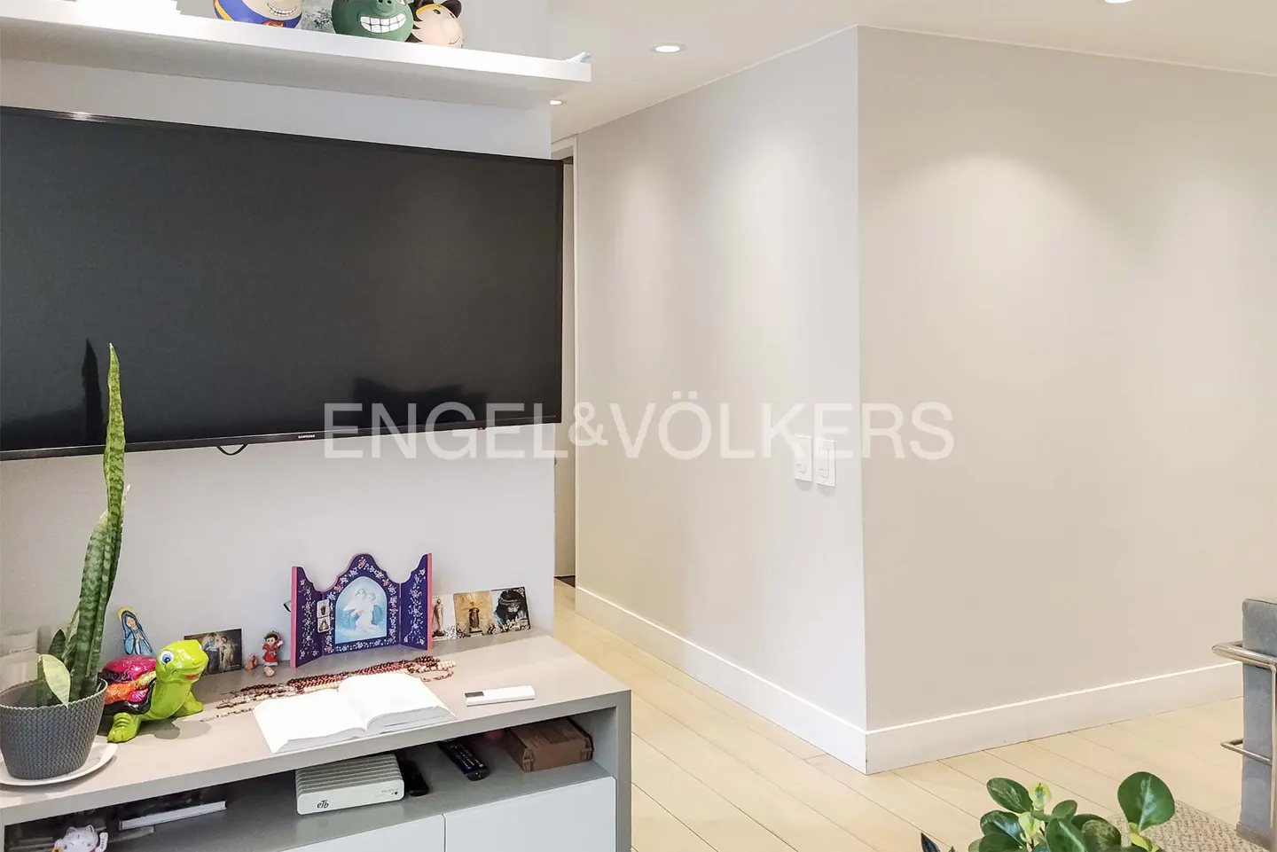 A wall-mounted TV above a grey console table with a snake plant, decorative items, and an open book in a bright room.