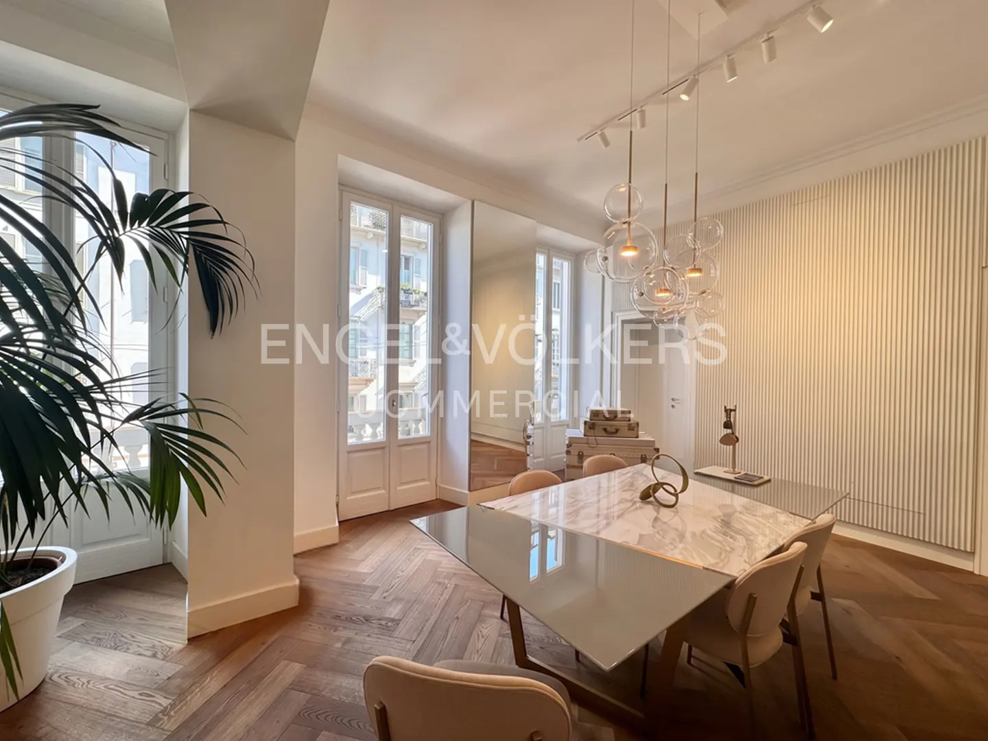 Bright dining room with herringbone wood floors, a marble-topped table, and bubble chandelier. White walls and a large potted plant add to the airy feel.