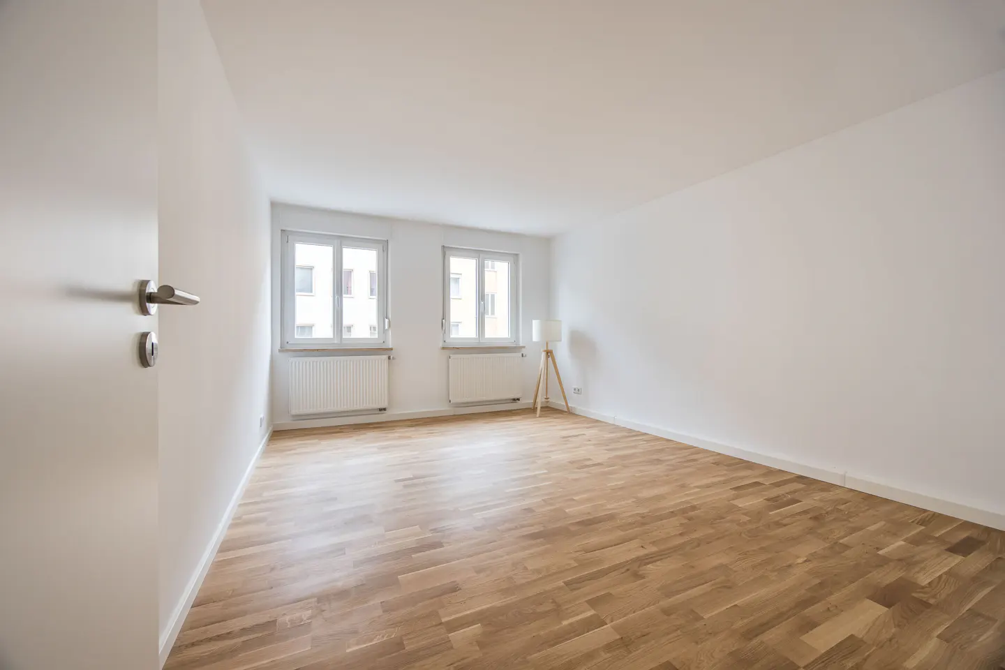 Bright, empty room with hardwood floors, white walls, and two windows. A white door is ajar on the left. A floor lamp stands in the corner.