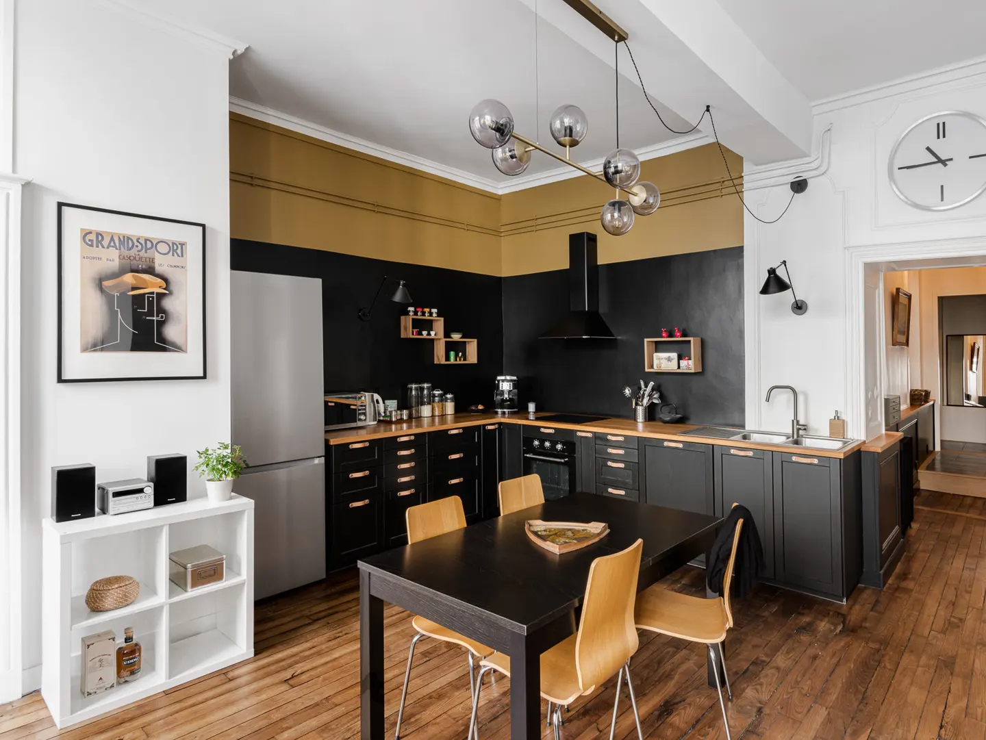 A modern kitchen with black cabinets, wood countertops, and a black dining table with four chairs. A stainless steel refrigerator and a white shelving unit are also visible.