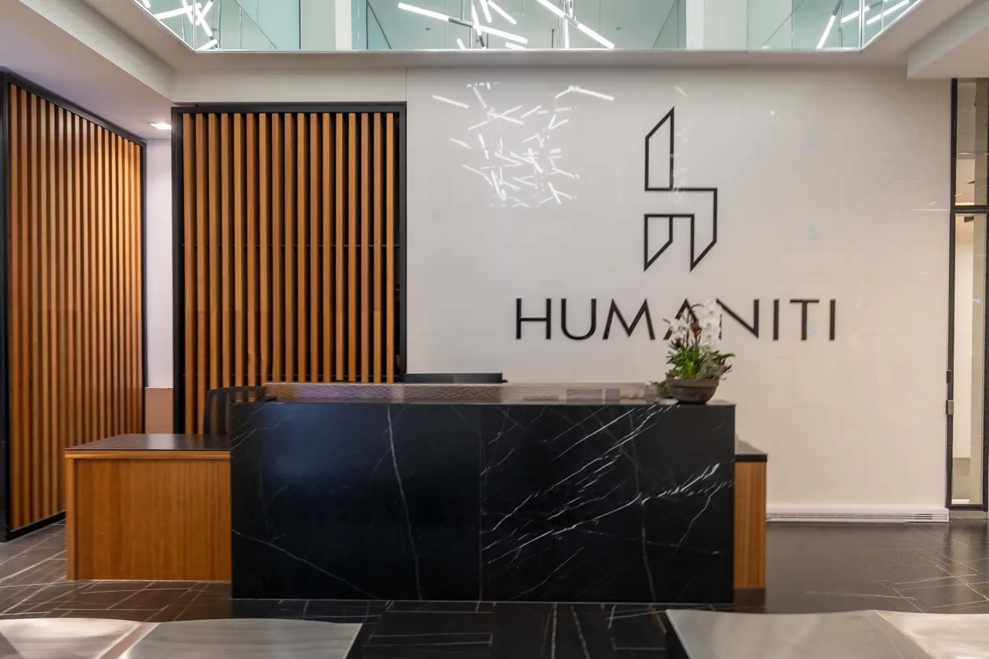 Modern office lobby with a black marble reception desk, wood slat walls, and the "Humaniti" logo on a white wall.