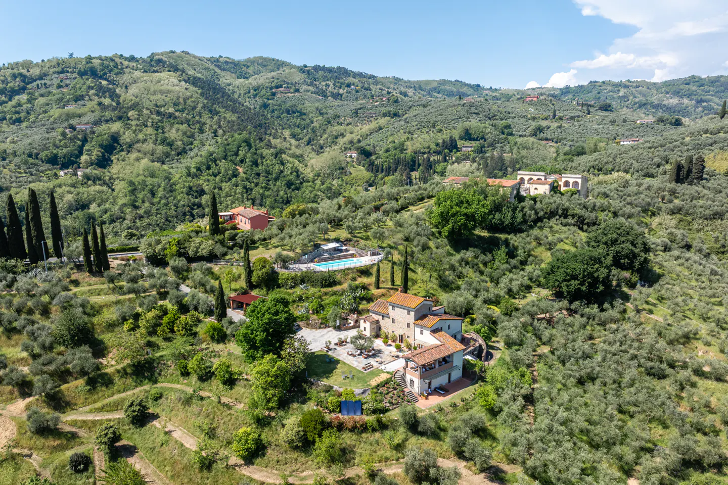 Aerial view of a Tuscan villa with a red tile roof, surrounded by olive trees and rolling green hills under a blue sky.