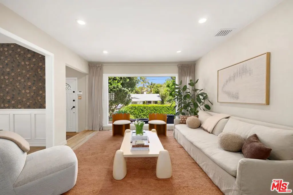 Living room with a long white sofa, two chairs, and a white coffee table on a brown carpet. A large window overlooks a green yard.