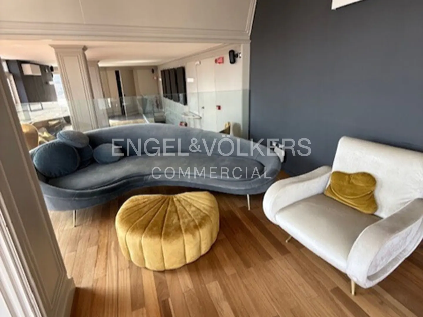 Interior view of a room with a blue sofa, gold ottoman, and white armchair on a wooden floor.