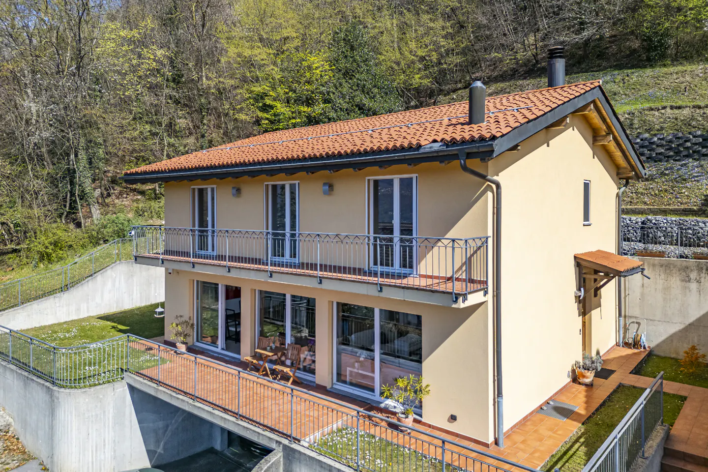 Two-story beige house with a red tile roof, black gutters, and balconies with metal railings, set against a green hillside.