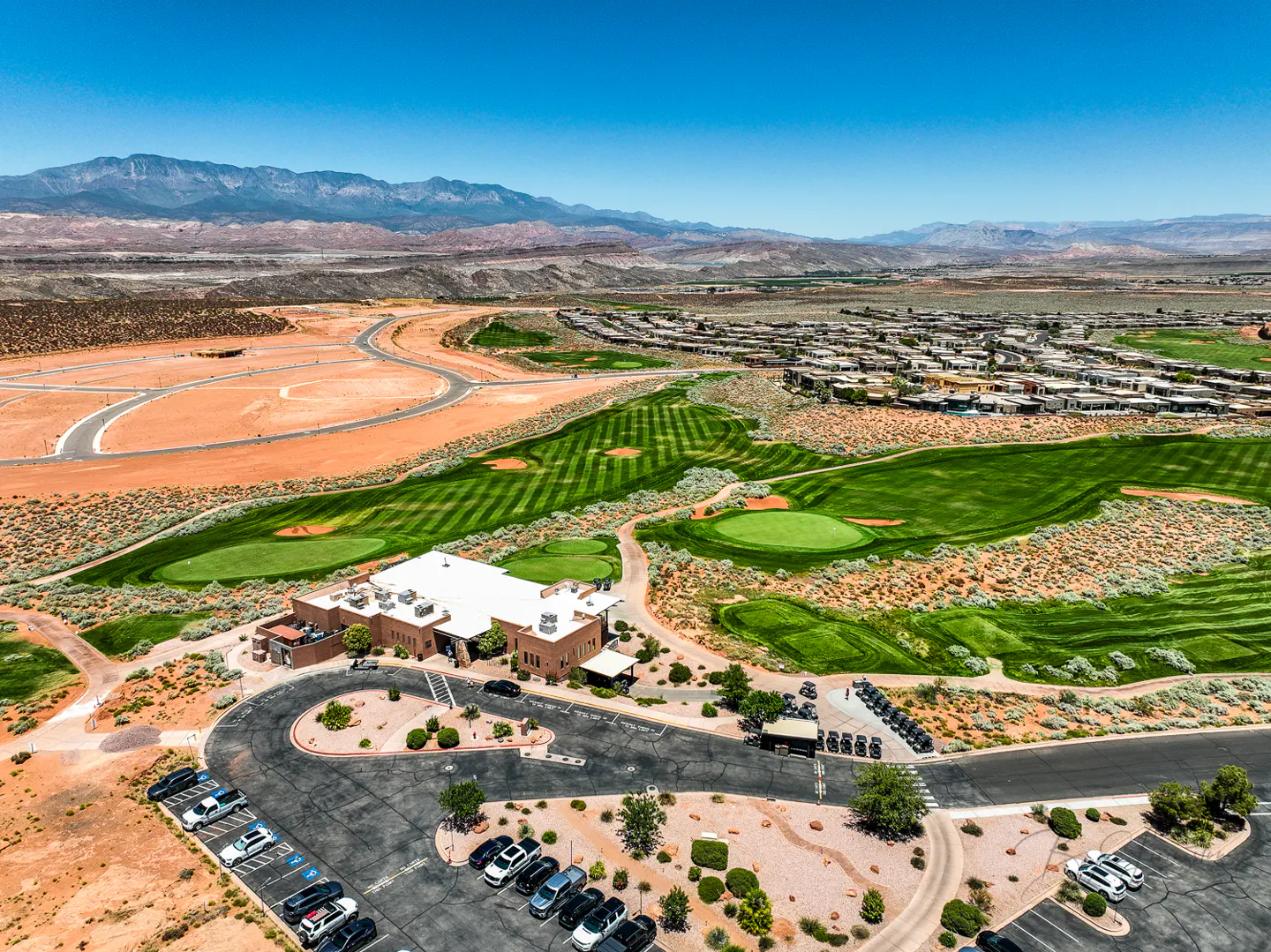 Aerial view of a golf course with a clubhouse, parking lot, and mountains in the background under a clear blue sky.