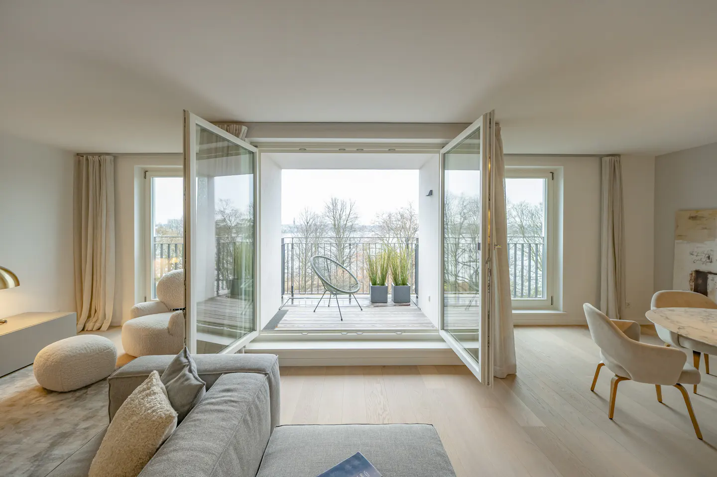 A bright living room with open doors to a balcony. A gray sofa and chairs are visible. The balcony has a chair and plants.