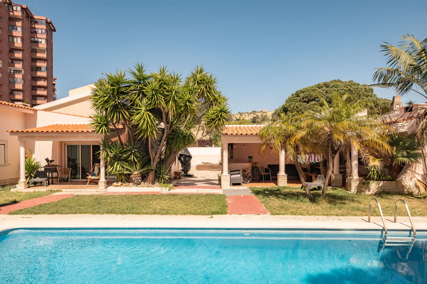 A sunny backyard with a turquoise pool, green lawn, and a peach-colored house with a red tile roof. Palm trees and a tall building are visible.