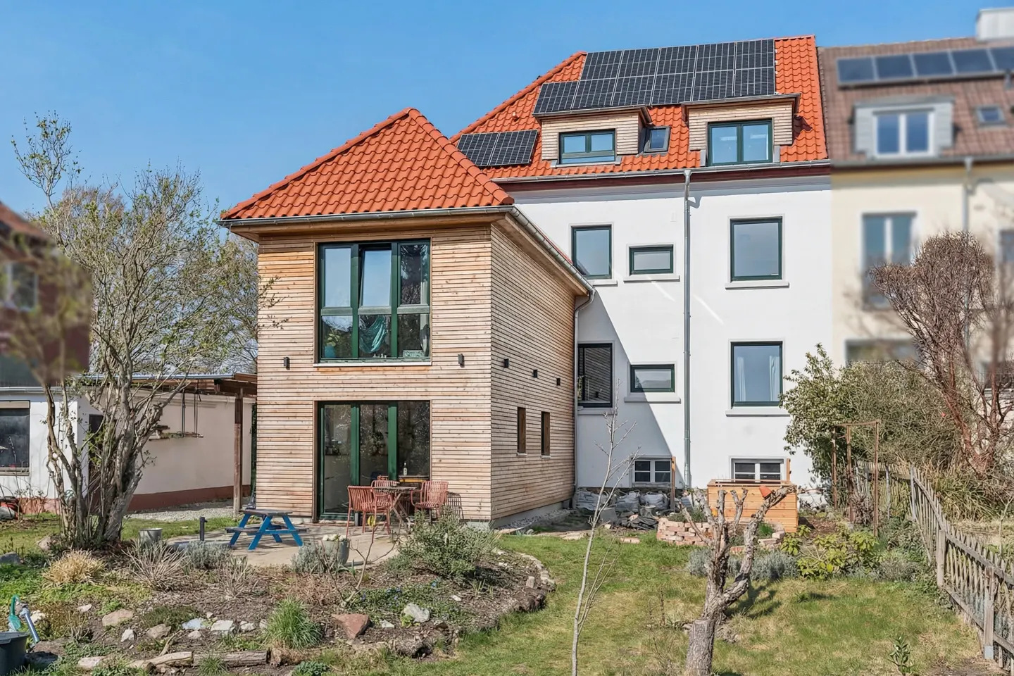 Exterior view of a modern house with a wood facade, red tile roof, and solar panels, surrounded by a green lawn and garden.