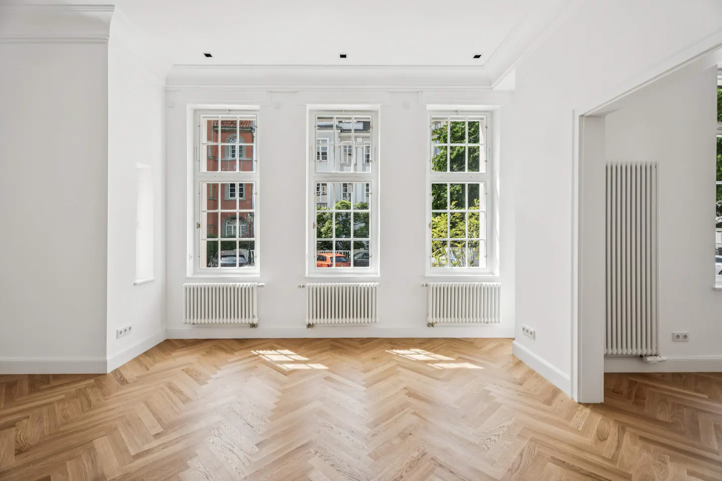 Bright, empty room with white walls, herringbone wood floors, and three large windows with white radiators underneath.