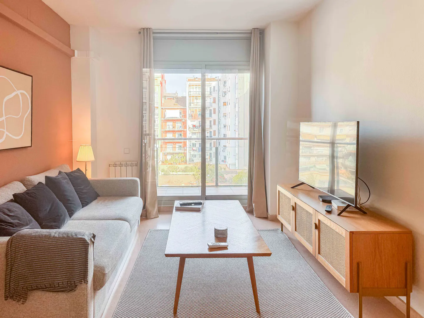 Bright living room with a gray sofa, wooden coffee table, and TV stand. Large window with city view. Abstract art on a peach wall.