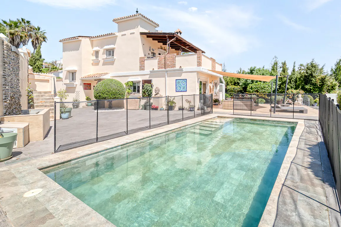 A tan two-story house with a pool, surrounded by a black fence, is set against a blue sky.