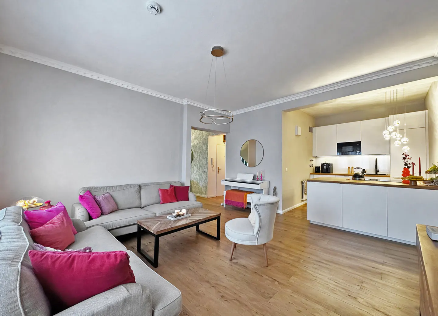 Open-concept living space with gray sofas, pink pillows, wood floors, and a white kitchen with modern appliances.