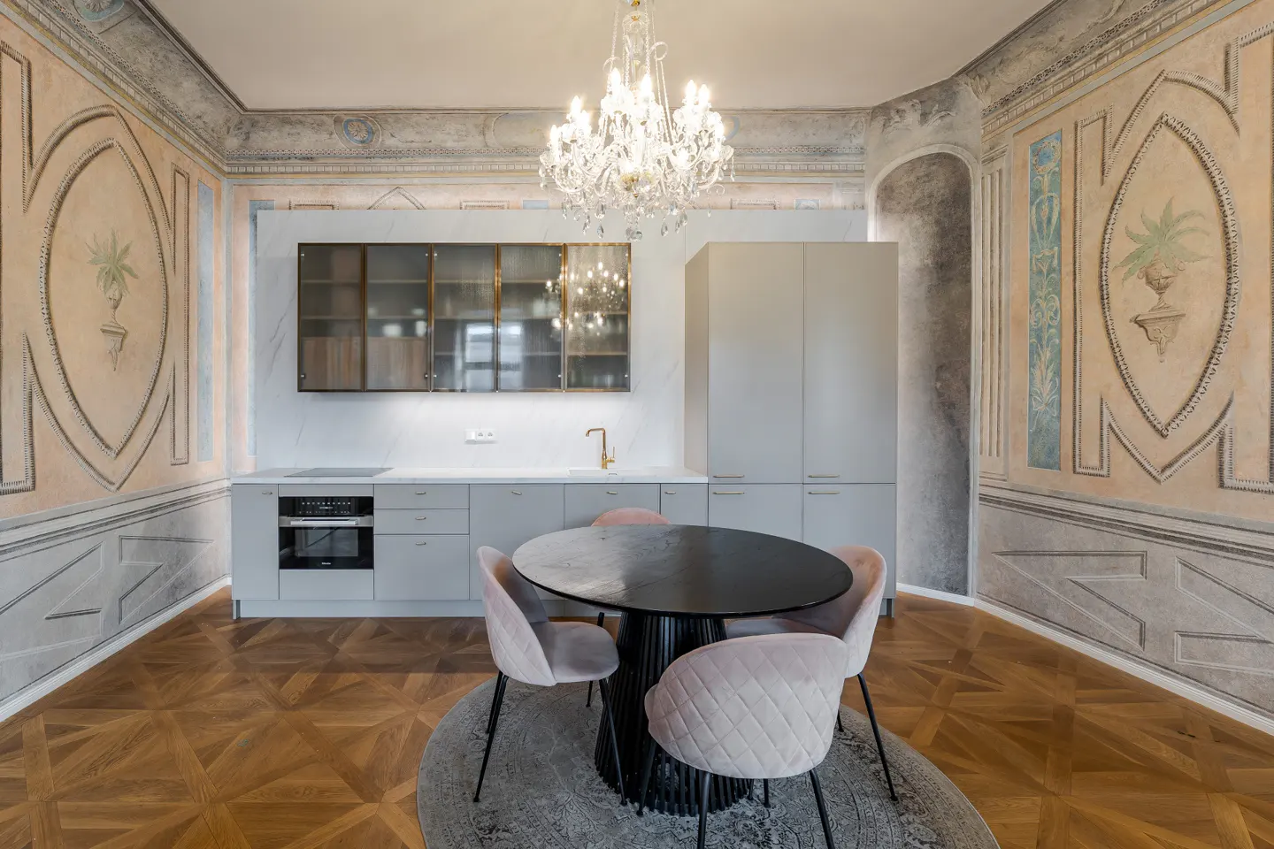 A modern kitchen with gray cabinets, a black table with pink chairs, and ornate wall paintings. A crystal chandelier hangs above the table.