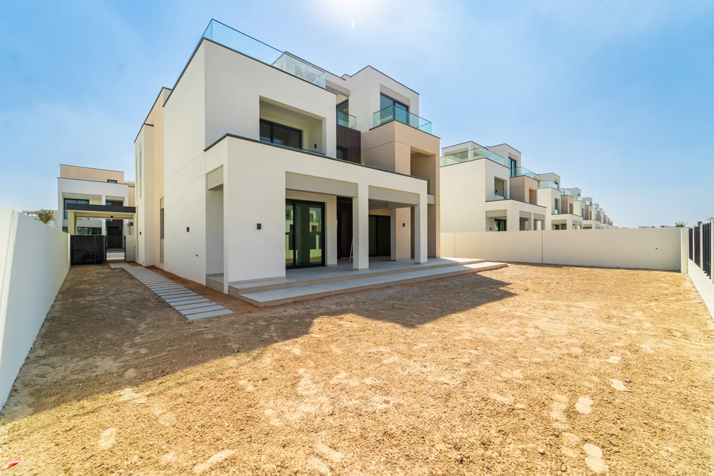 Modern two-story white house with a flat roof and glass balcony, set on a sandy lot under a clear blue sky.