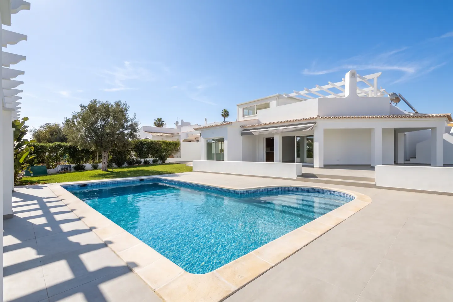 Modern white villa with a blue swimming pool, patio, and pergola under a clear blue sky.