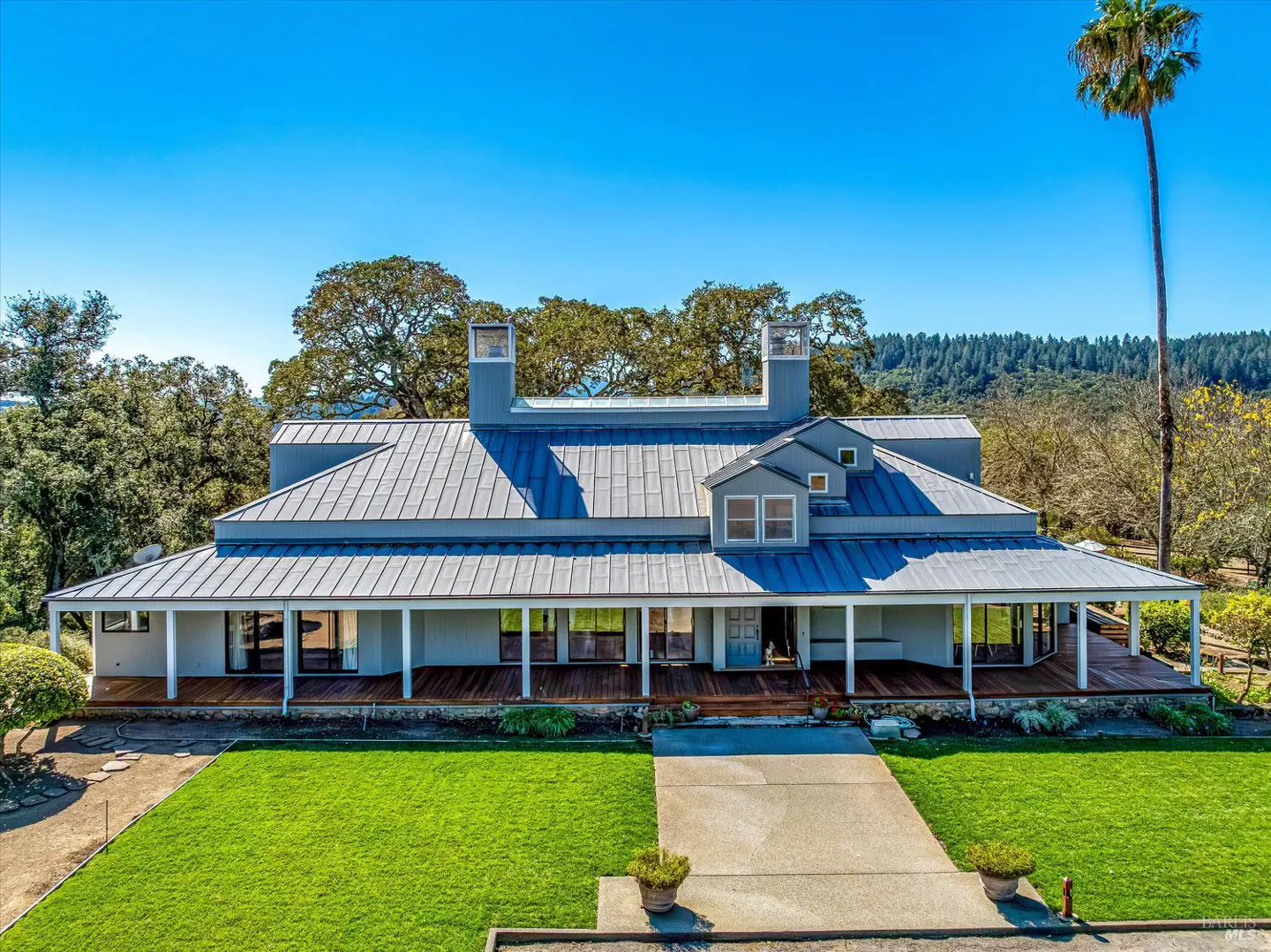 Aerial view of a gray farmhouse with a metal roof, a long porch, and a green lawn under a clear blue sky.
