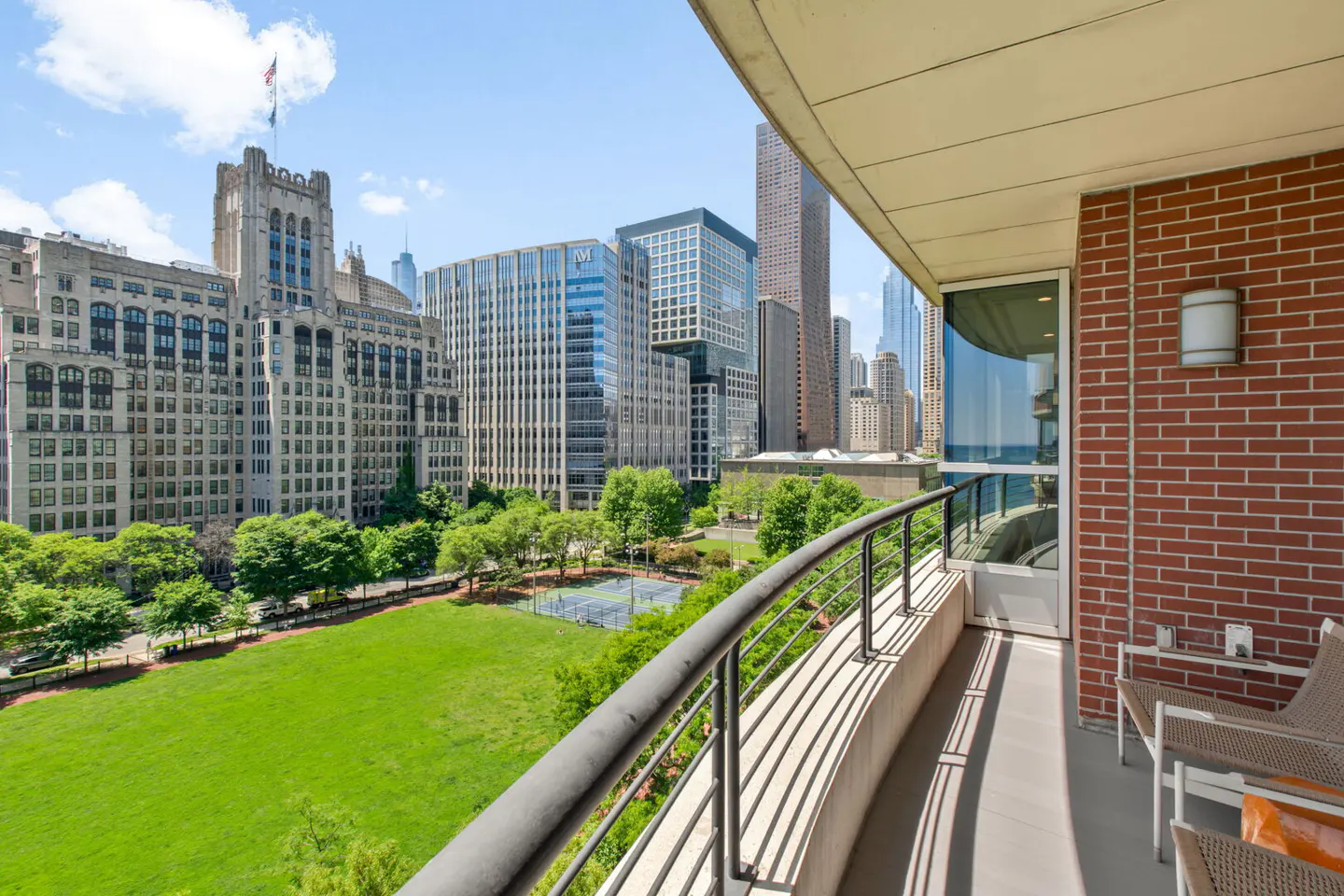 Balcony view overlooking a green park, tennis courts, and a city skyline with historic and modern buildings on a sunny day.