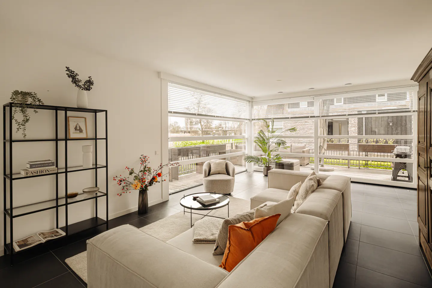 Bright living room with a beige sectional sofa, black tile floor, and large windows overlooking a patio. A black bookshelf stands against a white wall.