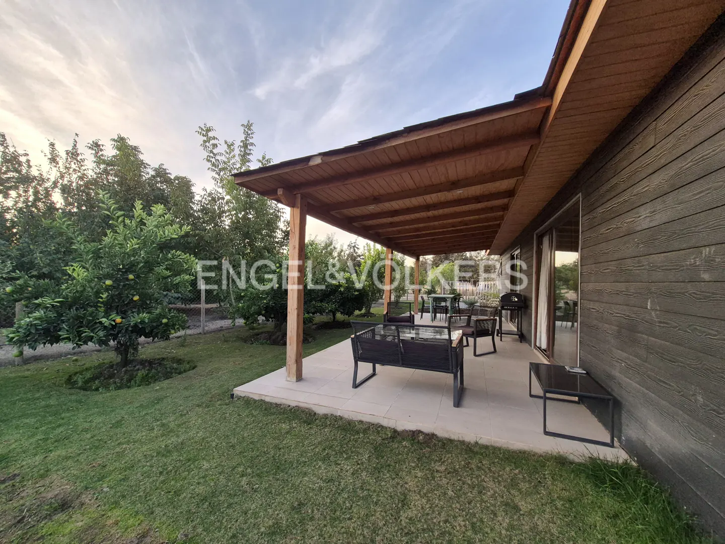 Exterior view of a house with a wooden patio, furniture, and a green lawn with trees.