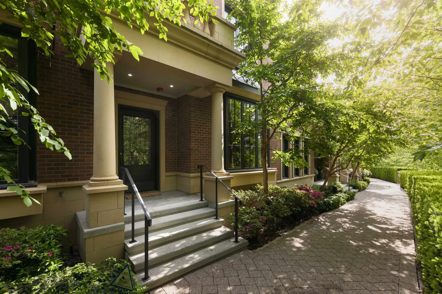 Brick house entrance with stone steps, black railings, and a black door. Lush green trees and bushes surround a stone path.
