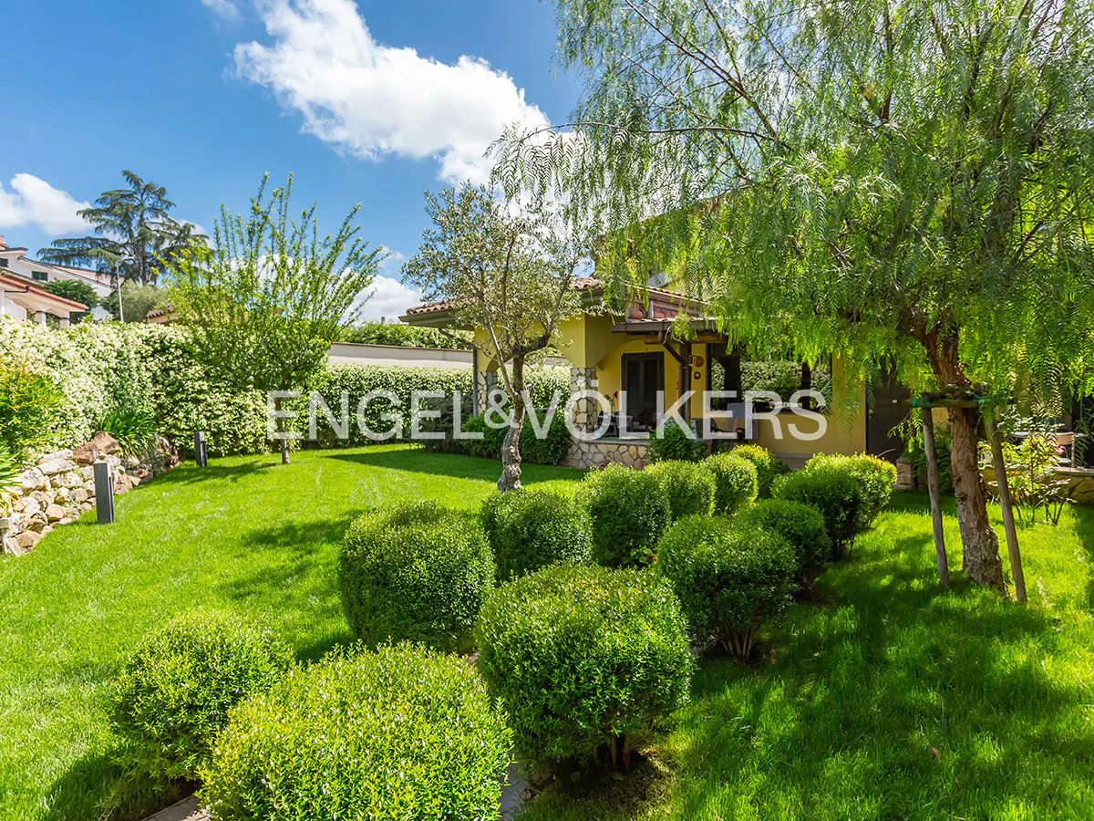 A yellow house with a green lawn and trees under a blue sky with white clouds. The Engel & Volkers logo is visible.
