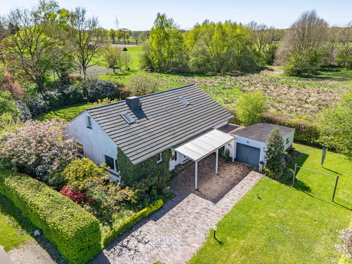 Aerial view of a white house with a gray roof, a carport, and a detached garage surrounded by green lawns and trees.