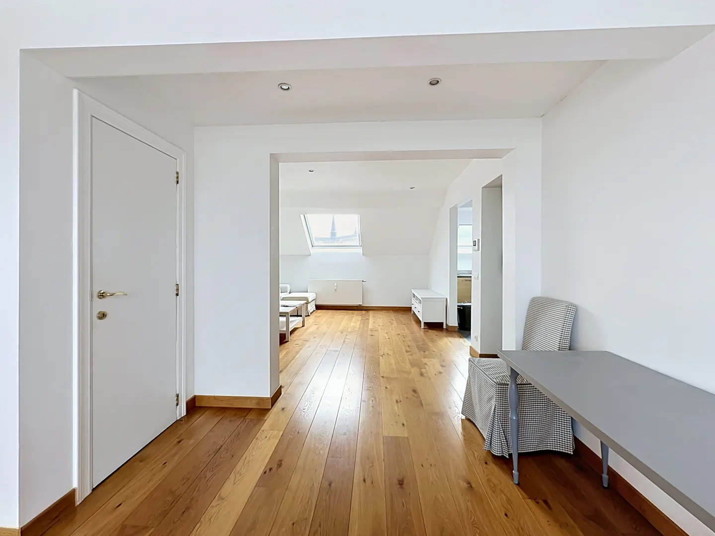 Bright, open apartment interior with hardwood floors, white walls, and a gray table with a covered chair. A doorway leads to a living area with a skylight.