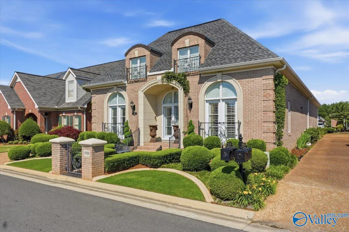 A tan brick house with a gray roof, arched windows, and manicured green bushes under a blue sky.