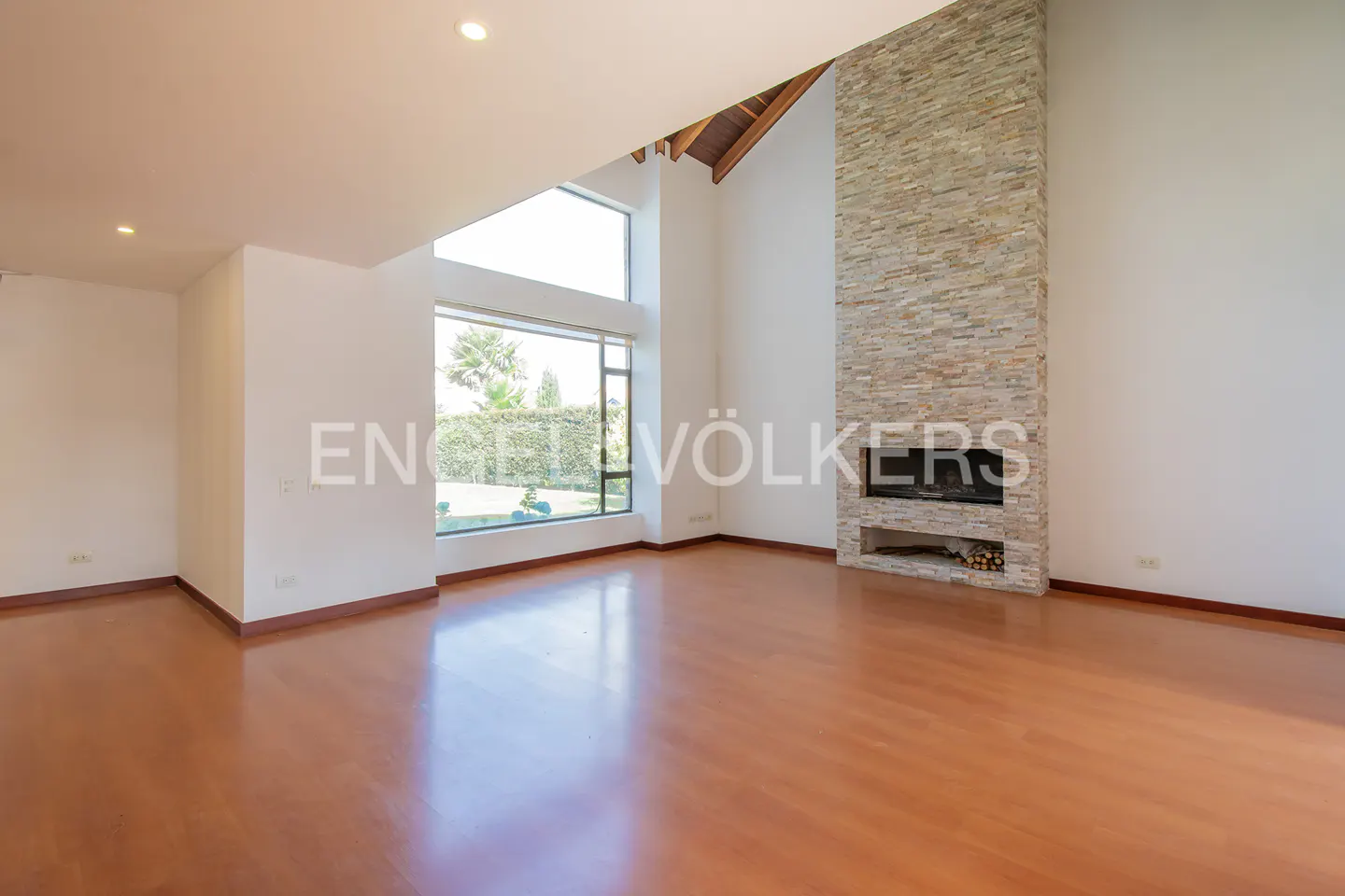Empty living room with hardwood floors, white walls, and a stone fireplace. A large window shows a green lawn.