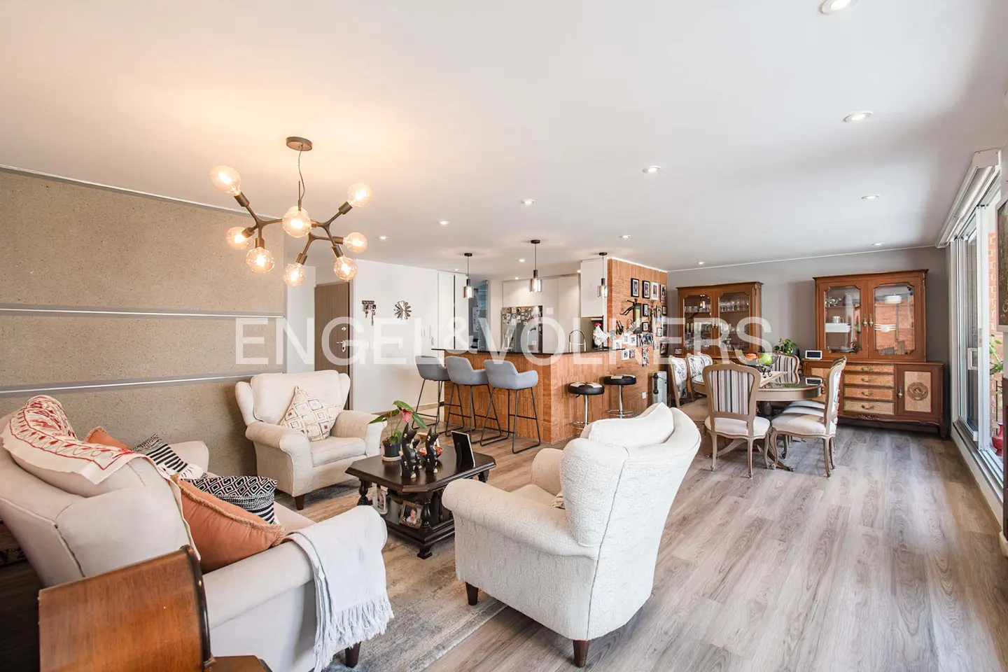 Open-concept living space with white sofas, a wood-paneled kitchen island with bar stools, and a dining area with wooden cabinets.
