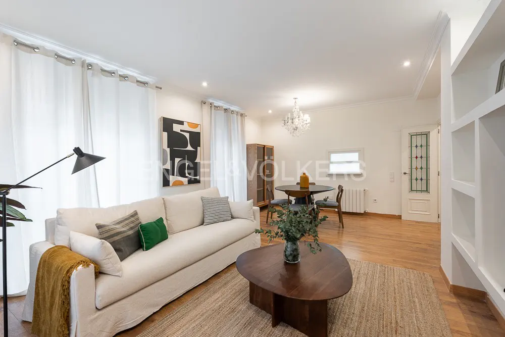 Bright living room with white sofa, wood floors, and a dining area with a chandelier. A modern painting hangs on the wall.
