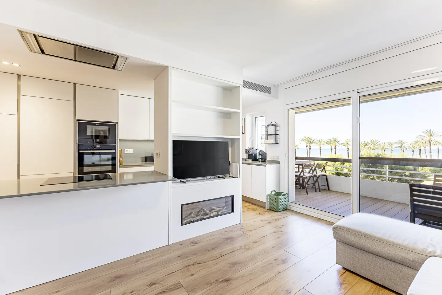 Bright, modern apartment interior with white cabinets, wood floors, and a balcony overlooking palm trees and the ocean.