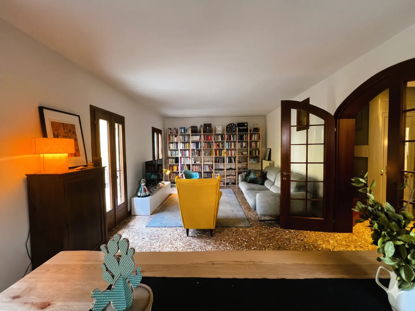 A bright living room with a large bookshelf, yellow armchair, and gray sofa. French doors lead to another room.