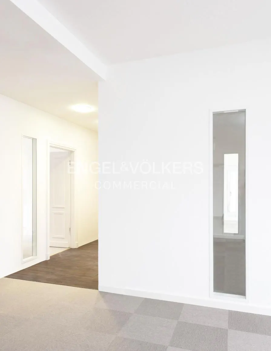 Bright, empty office space with white walls, gray carpet, and glass-paneled doors leading to other rooms. Engel & Völkers logo visible.