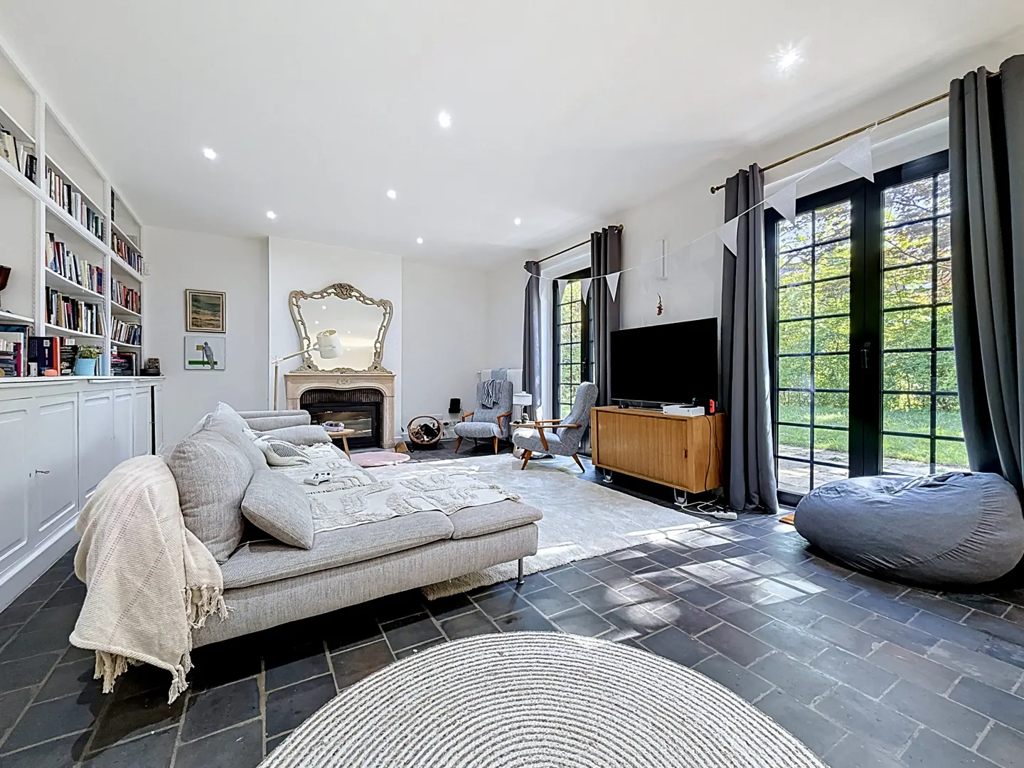 Bright living room with gray sofa, fireplace, and built-in bookcase. Black-framed doors lead to a green garden.