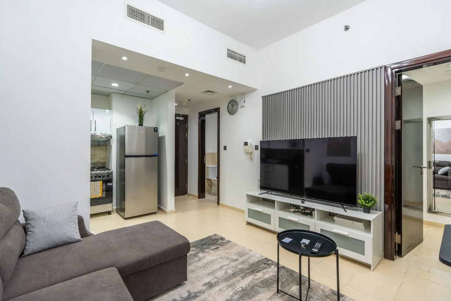 Apartment living room with a gray sofa, TV, and stainless steel refrigerator. The walls are white, and the floor is light beige.