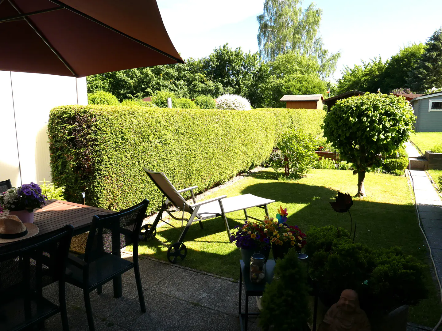 A sunny backyard patio with a dining set, a lounge chair on the lawn, a tall hedge, and a small tree under a blue sky.